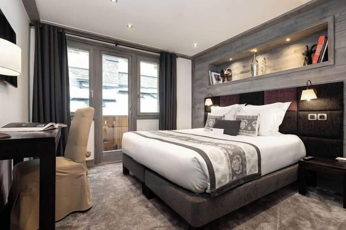Una habitación de hotel moderna con una cama grande, un escritorio y una silla. Se ve una ventana con cortinas, una estantería con objetos decorativos y una mesita de noche con una lámpara.