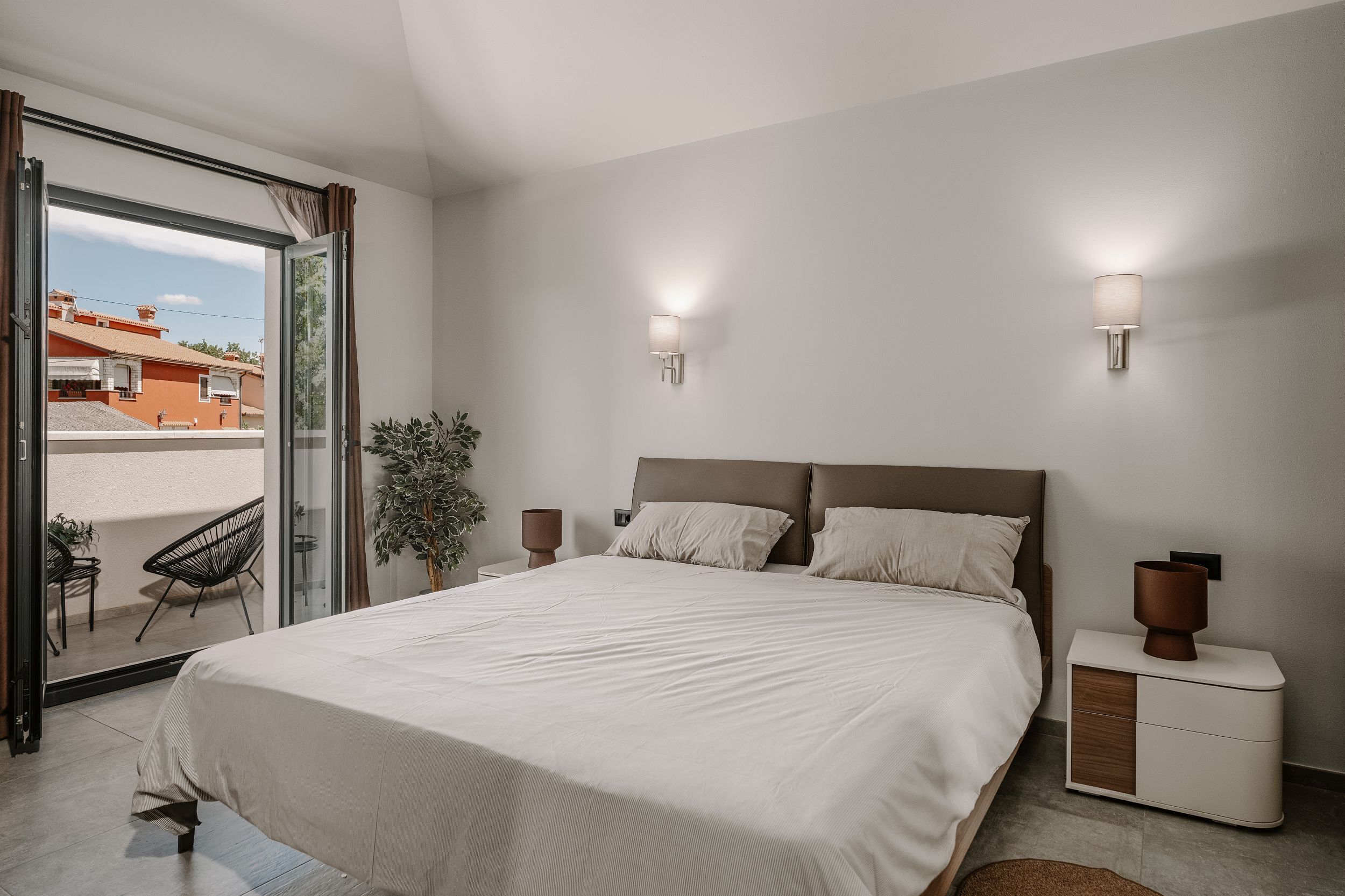 Ein Schlafzimmer mit einem großen Bett, einem Nachttisch, einer Zimmerpflanze und Blick durch eine Schiebetür nach draußen.
