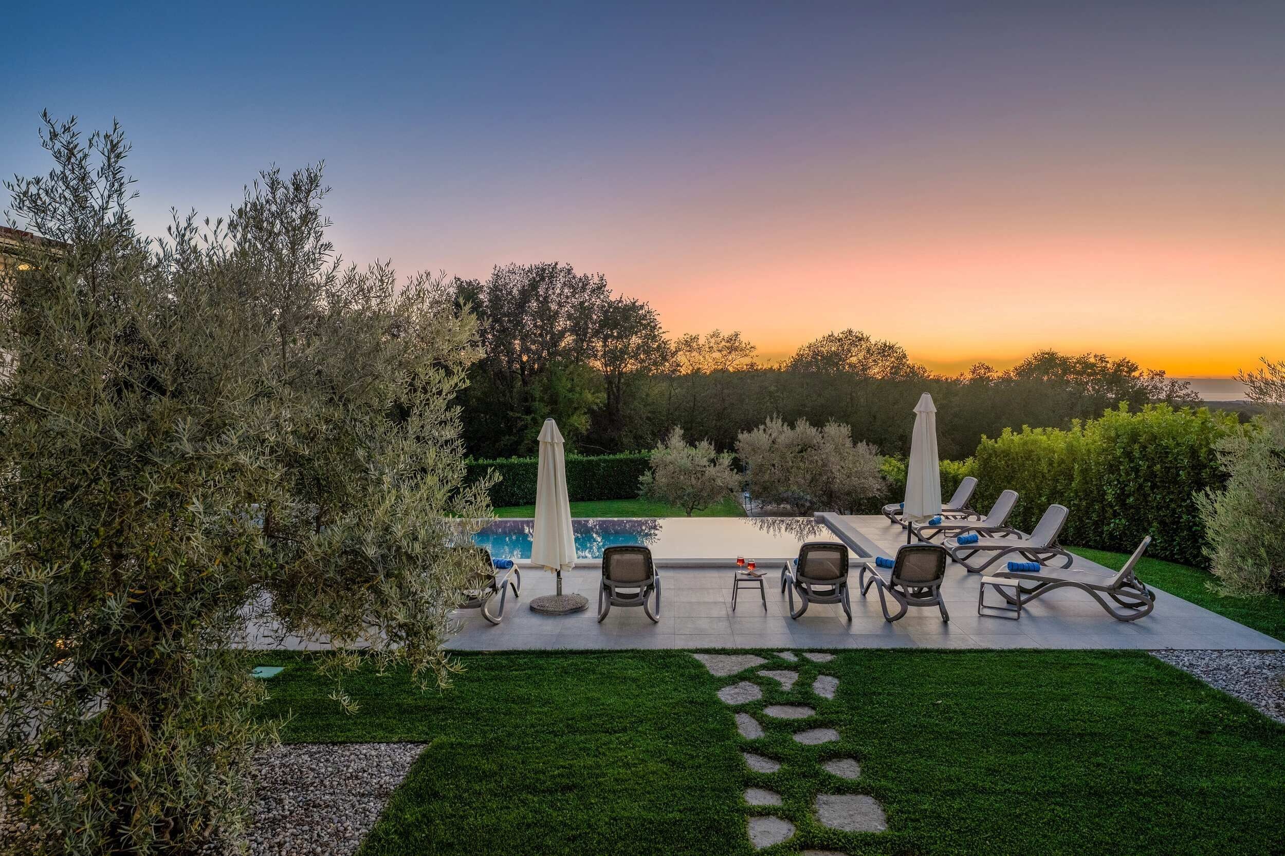 A poolside area with lounge chairs and umbrellas, surrounded by greenery and a view of the sunset.