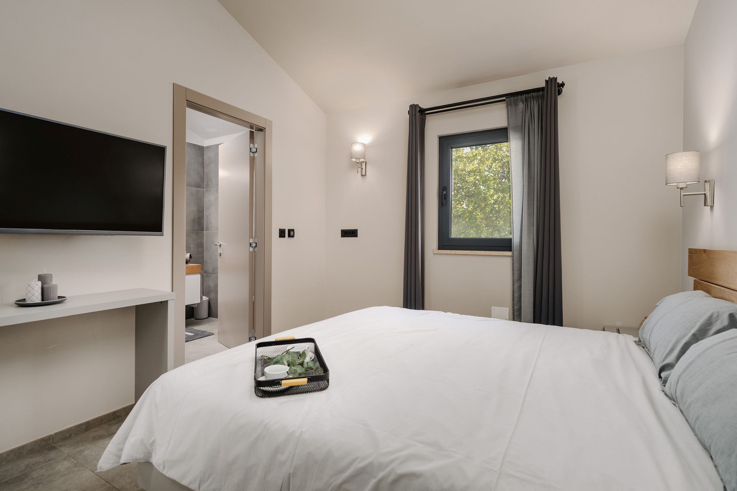 A modern hotel room with a large bed, a flat-screen TV mounted on the wall, a window with curtains, and a small table with a tray of items.