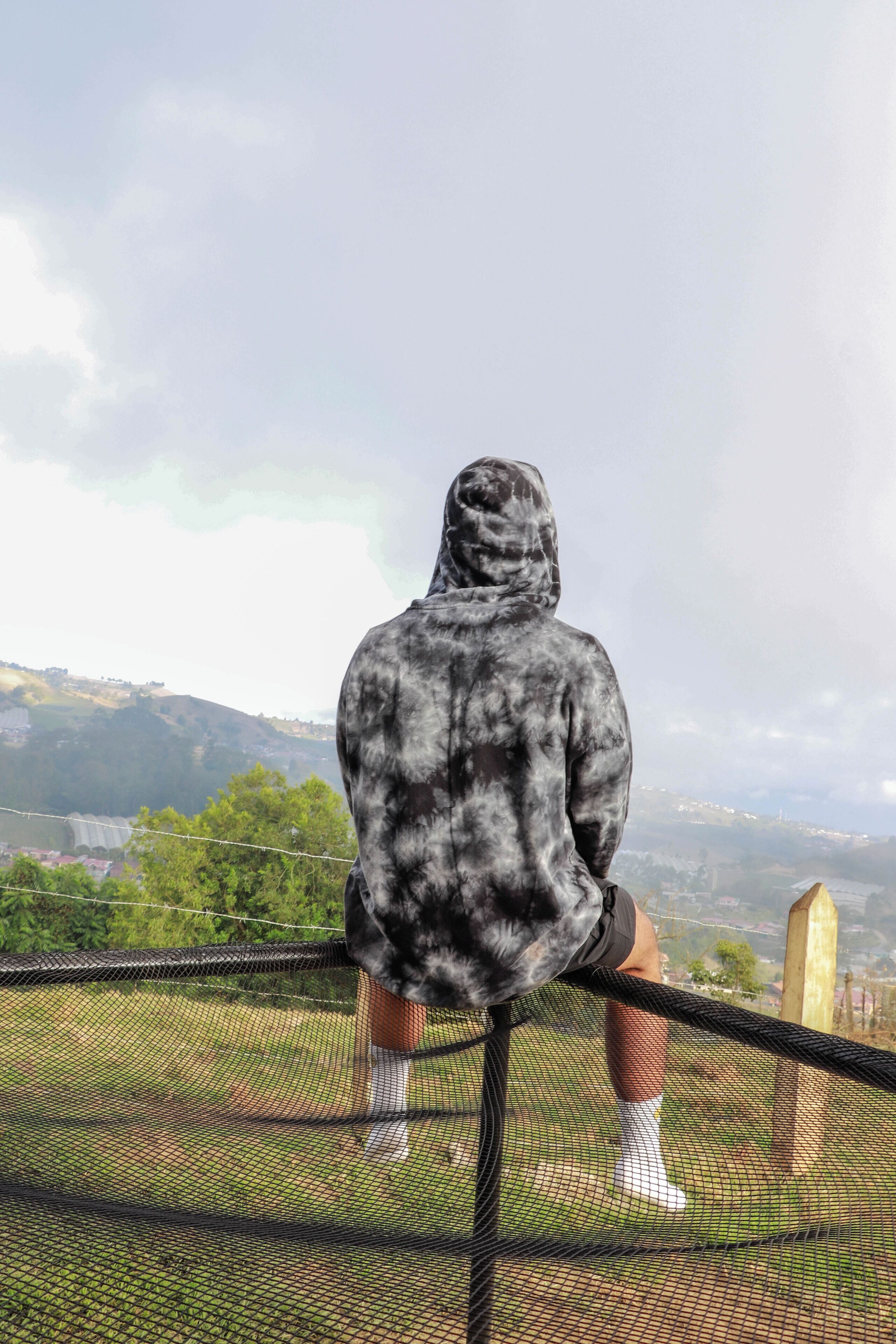 A person sitting on a metal railing overlooking a scenic landscape with hills and a body of water.