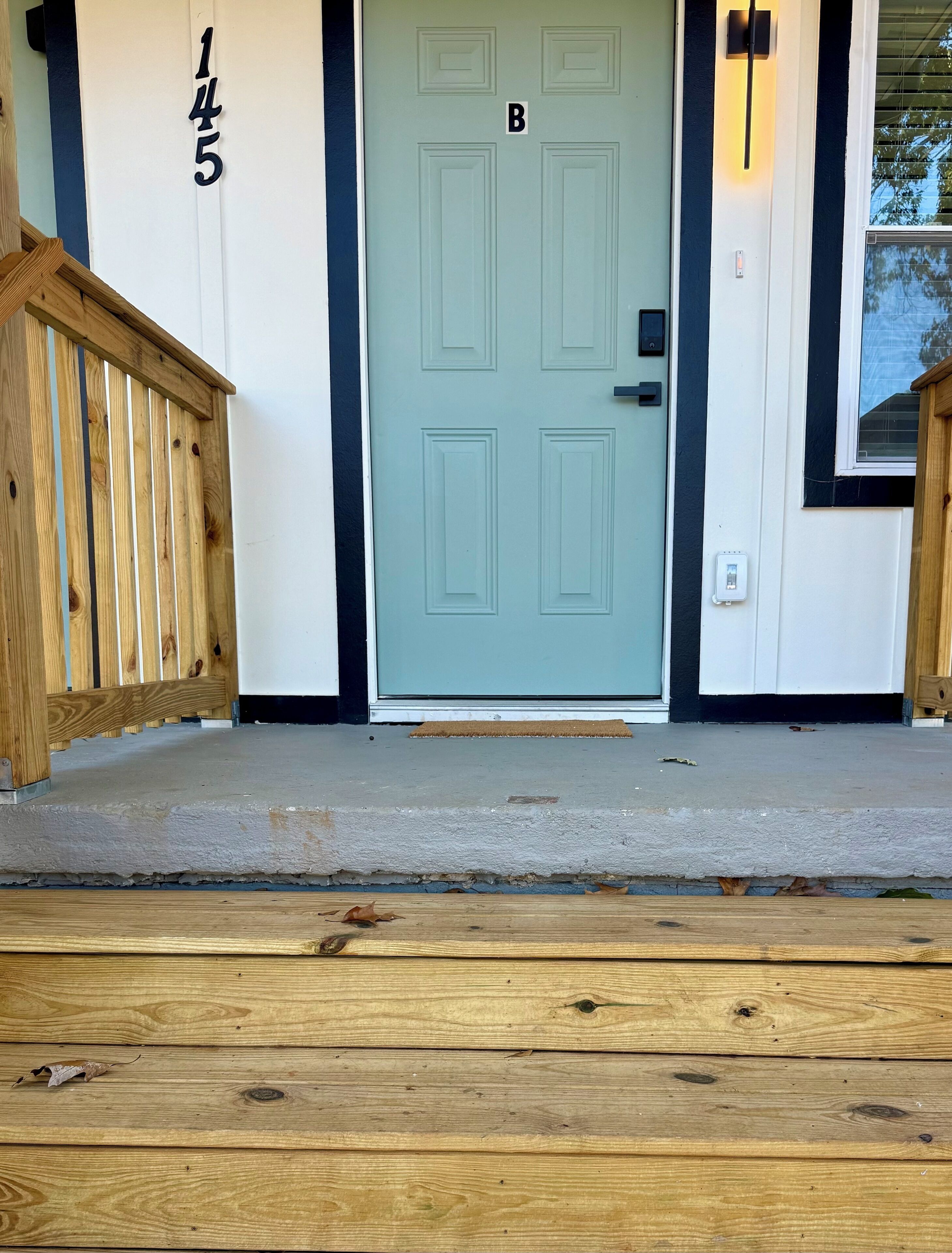 Une porte d’entrée avec le numéro « 145 » et la lettre « B » au-dessus, une rambarde en bois et un porche en béton.
