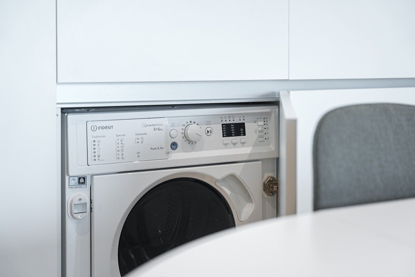 A front view of a washing machine with control panel, water inlet, and a grey chair in the foreground.