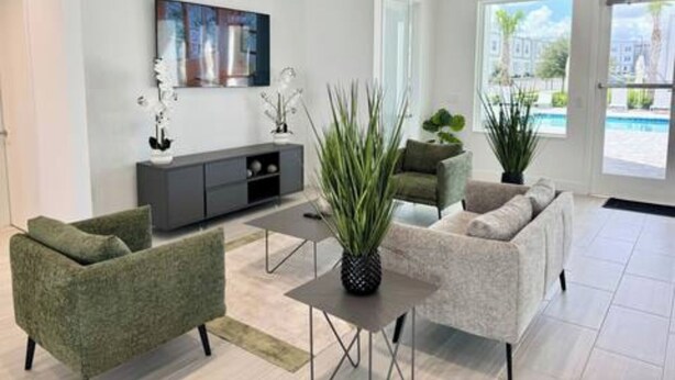 A modern living room with a grey sofa set, a coffee table, and a TV mounted on the wall. There are potted plants and a view of a pool through the door.
