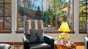 Quarto com uma janela grande que oferece vista para árvores e um edifício, uma poltrona de couro com almofada estampada e uma mesa lateral de madeira com luminária.