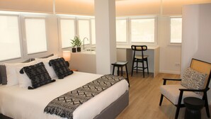 Una habitación de hotel moderna con una cama grande, un escritorio y una silla. Se observan ventanas con persianas, una planta en maceta y una lámpara sobre el escritorio.