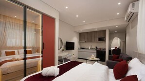 Une chambre d’hôtel moderne dotée d’un grand lit, d’un coin salon séparé, d’une kitchenette et d’une porte-fenêtre coulissante donnant sur un balcon.