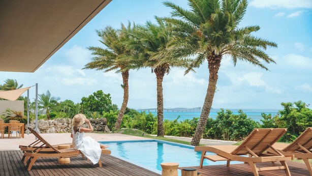 Terraza de madera con sillones de descanso y piscina, rodeada de palmeras y vista al océano.