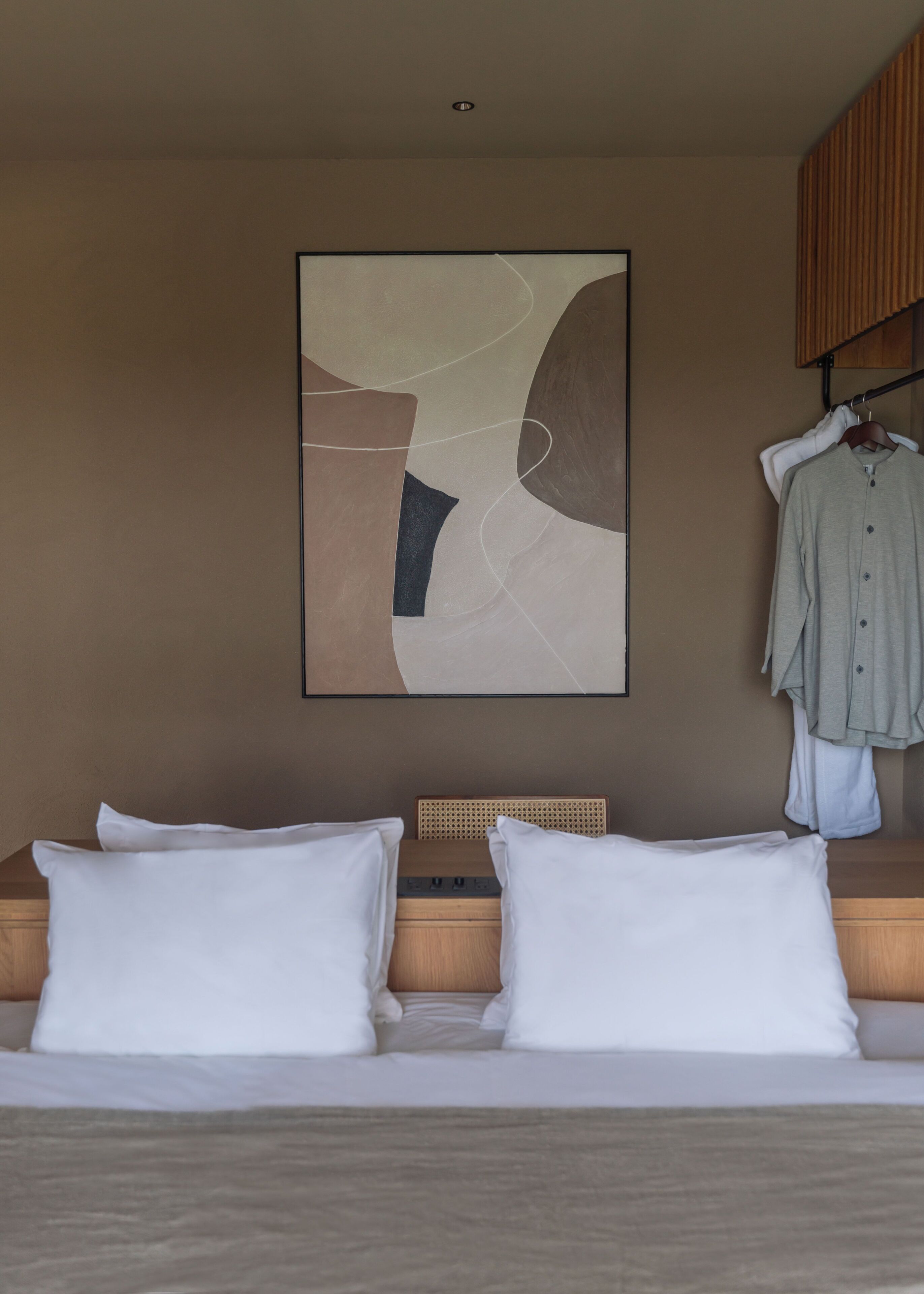 Hotelkamer met een bed, kussens, een hoofdeinde, een wanddecoratie in abstractie en een kledingrek met kleding.