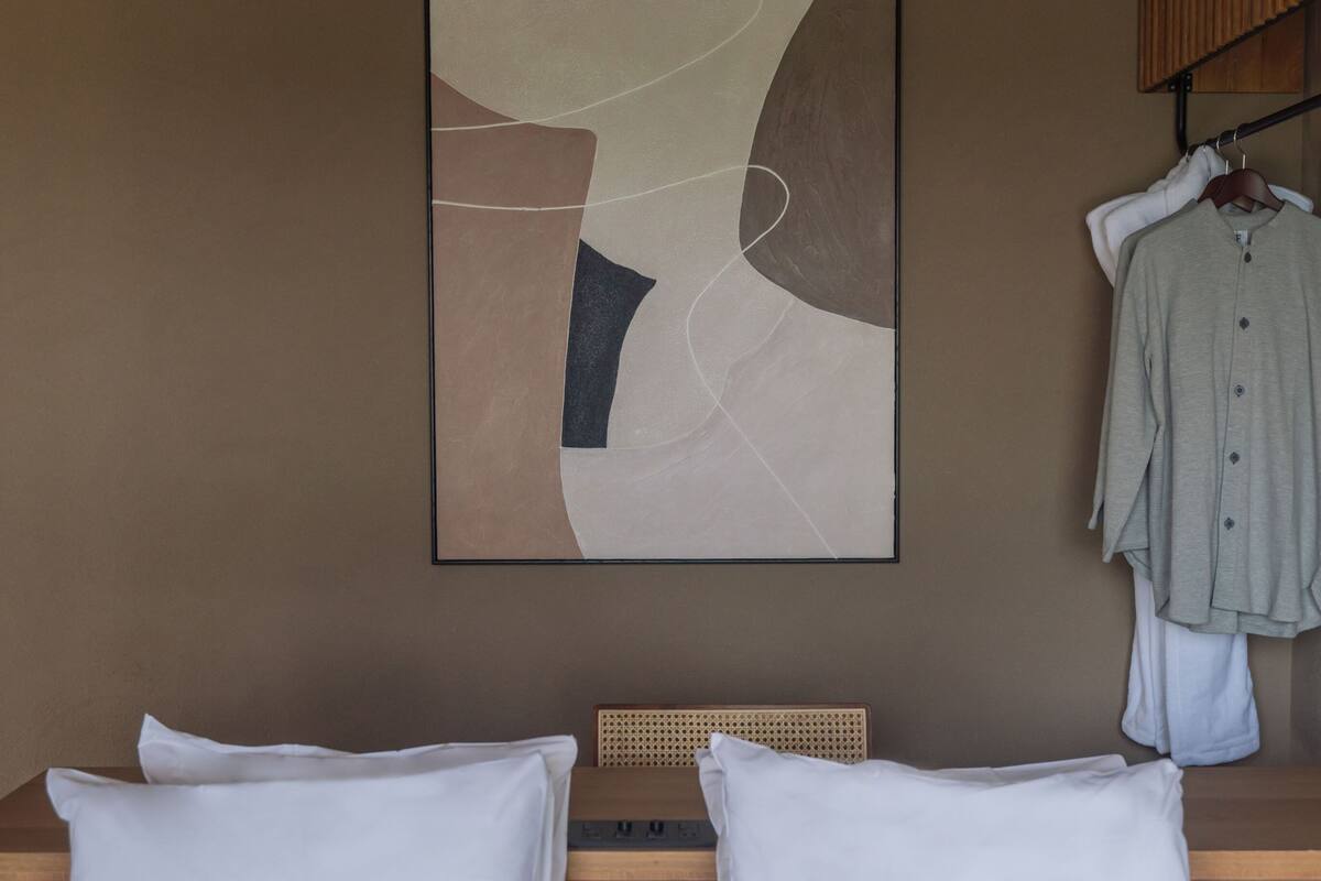 Hotellihuone, jossa on sänky, tyynyt, sängynpääty, seinälle ripustettu abstrakti taulu ja vaateteline vaatteineen.