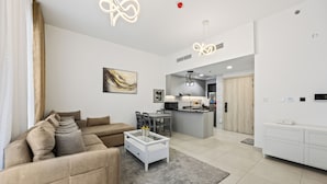 A modern living room with a beige sofa, a white coffee table, and a kitchen area in the background. - Calm Balcony Apartment with Pool Access (Dubai)
