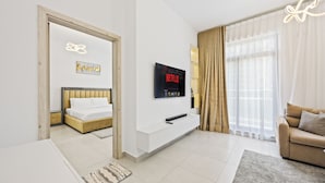 A modern hotel room with a bed, a TV mounted on the wall, a sofa, and a large window with curtains. - Calm Balcony Apartment with Pool Access (Dubai)