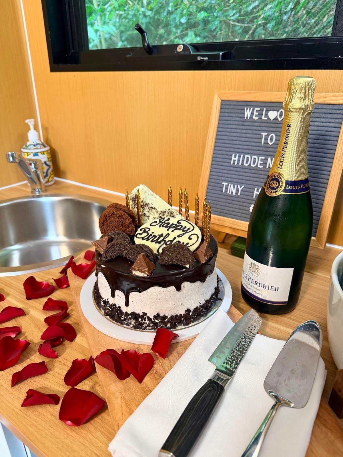 A birthday cake with chocolate and white frosting, a bottle of Louis Perrier champagne, and a sign that says 'WELCOME TO HIDDEN NINY H.'