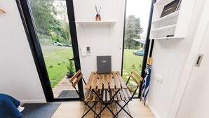 Une pièce de taille modeste, avec une table et des bancs en bois, une petite étagère sur laquelle repose un livre, et une vue sur un jardin par une porte ouverte.