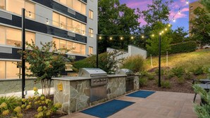 An outdoor patio with a stone barbecue and grill station, surrounded by a landscaped garden and a building in the background. - Elevated Minimalist Escape, Free Parking & Metro (Arlington)
