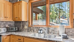 A kitchen with wooden cabinets, granite countertops, a built-in toaster, a sink, and a window overlooking a forested area. - Breckenridge Basecamp | Hot Tub + Fire Pit (Breckenridge)