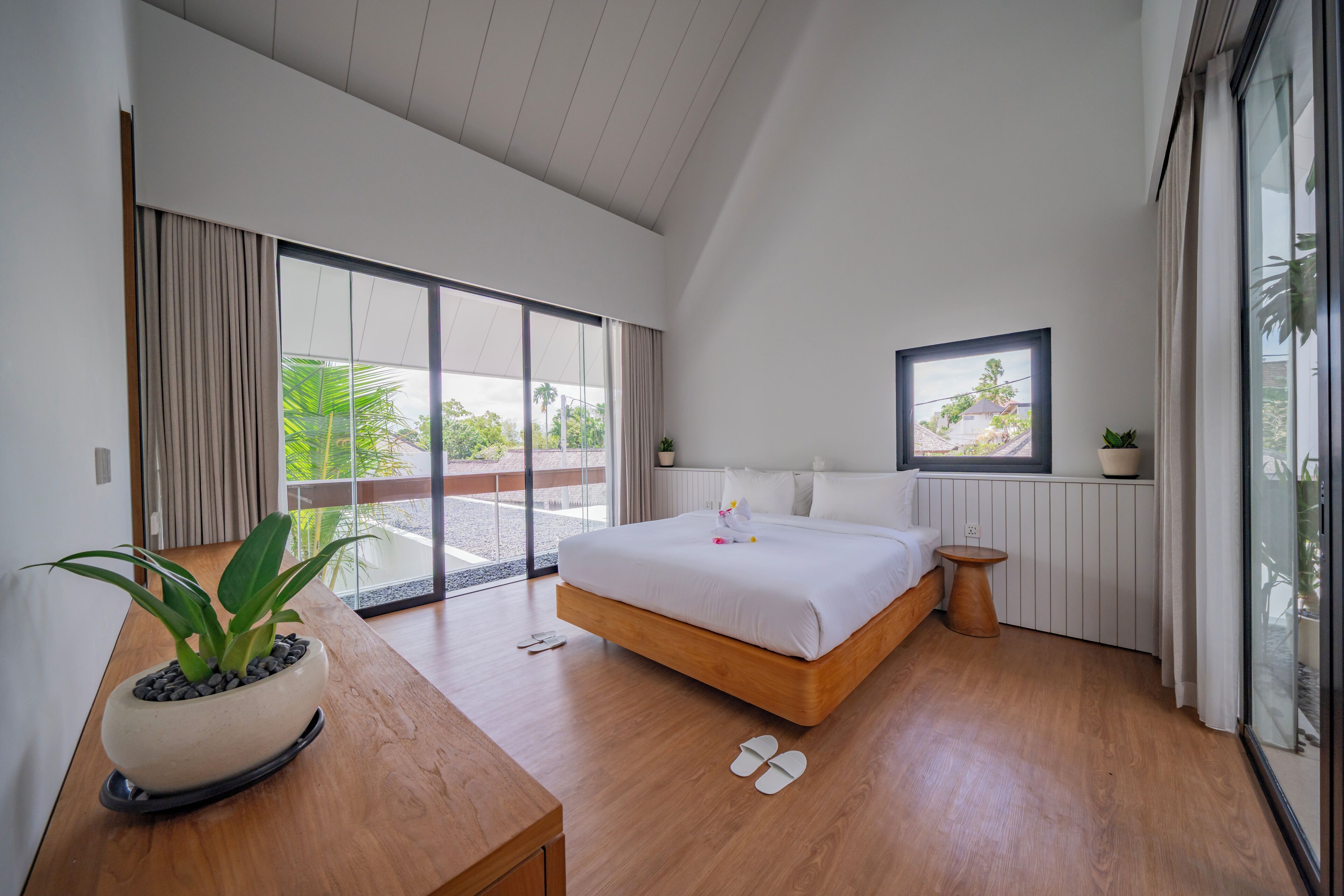 A modern bedroom with a large bed, a wooden nightstand, a potted plant, and a view of the outdoors through a sliding glass door.