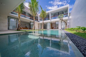 Un área de piscina moderna con agua cristalina, rodeada de palmeras y un edificio de varios pisos con amplios ventanales.