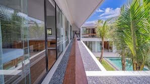 A modern, glass-walled corridor with a wooden walkway, leading to a pool area with palm trees. - Villa Shanaya Pererenan (Canggu)