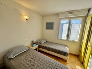 A small, single-room accommodation with two single beds, a bedside table with a lamp, a window with curtains, and a wall-mounted air conditioning unit.