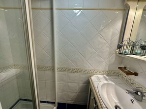 A bathroom with a glass shower enclosure, a white sink, and a shelf with toiletries.