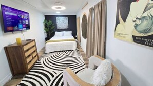 Una moderna camera d'albergo con un letto, una TV fissata al muro, una cassettiera in legno, un tappeto a motivi zebrati e un quadro appeso al muro.