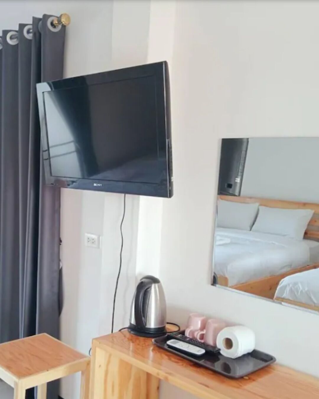 Ett hotellrum med en väggmonterad platt-TV, ett litet träbord med en vattenkokare, en fjärrkontroll och en toalettpappersrulle.