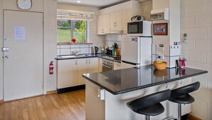 A modern kitchen with white cabinets, a central island with bar stools, a microwave, and a clock on the wall. - SOHO Vibes - 2 Bedroom Unit (South Hobart)