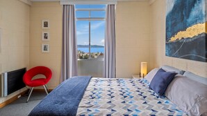 A hotel room with a bed, a red chair, a painting, and a view of the city. - SOHO Vibes - 2 Bedroom Unit (South Hobart)