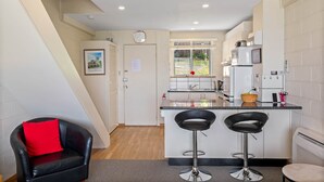 A compact kitchen with a central island, two bar stools, a red pillow on a chair, and a window with a view of greenery. - SOHO Vibes - 2 Bedroom Unit (South Hobart)