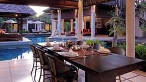 Un espace repas avec vue sur la piscine, comprenant une longue table dressée pour le dîner, entourée de chaises, et un coin salon couvert avec des coussins.