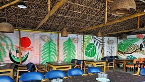 Un restaurant avec des plafonds en bambou, des fresques murales représentant des feuillages verts et des tables en bois agrémentées de coussins bleus.