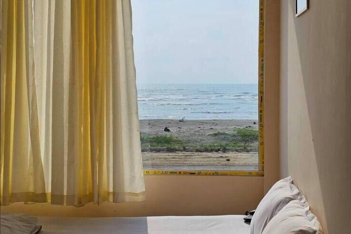 Ein Zimmer mit Bett, einem Fenster mit gelben Vorhängen und Blick auf den Strand.