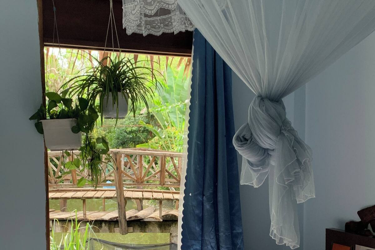 Une chambre avec un lit, une fenêtre donnant sur un paysage verdoyant et une moustiquaire suspendue.