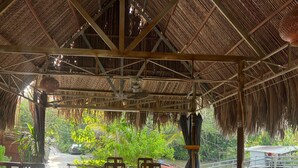 An open-air dining area with wooden tables and chairs, a thatched roof, and a view of greenery and water. - Mương Đình Lodge (Can Tho)