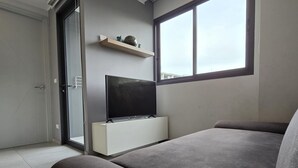 Una habitación moderna con un sofá gris, un televisor de pantalla plana sobre un soporte blanco, un ventanal corredero y una ventana con vistas.