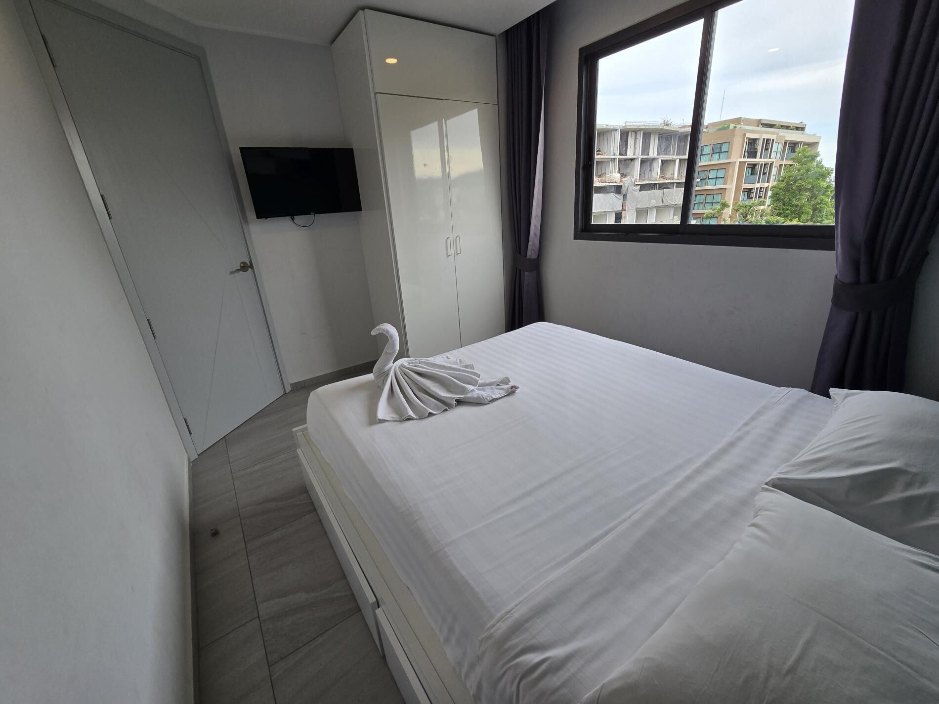 Habitación de hotel con cama, armario, televisor y ventana con vista a edificios.