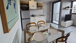 Una cocina funcional con refrigerador, microondas y estufa. Hay una mesa de comedor con sillas y superficie de vidrio. Un televisor está montado en la pared.