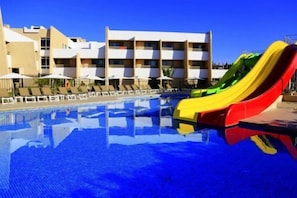 A swimming pool with a multi-colored slide, lounge chairs, and a building with balconies in the background.