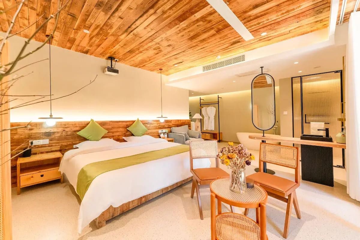 Quarto de hotel moderno com teto de madeira, uma cama grande com roupa de cama verde e branca, uma mesinha de cabeceira, uma cadeira, uma pequena mesa redonda com um vaso de flores e um espelho sobre a pia.