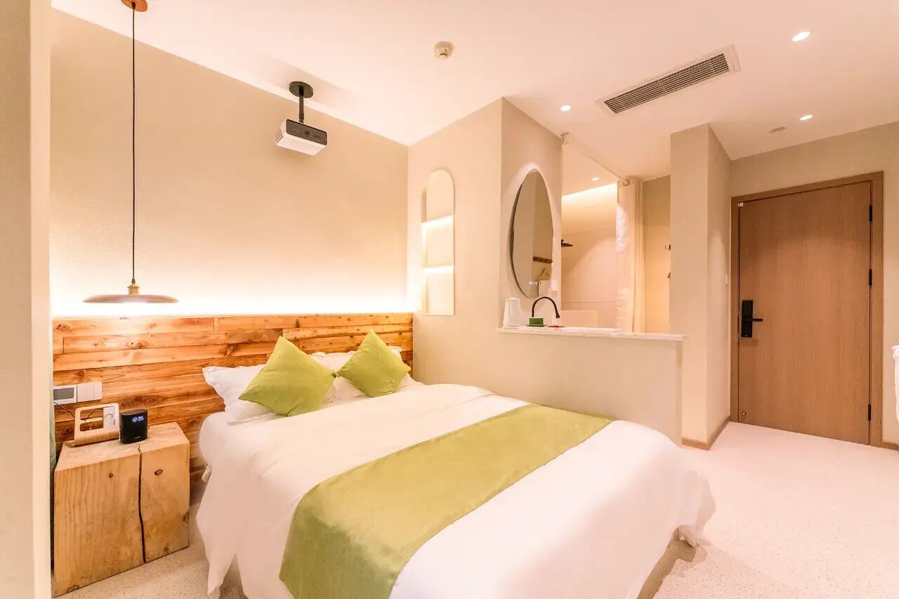 Un dormitorio con cabecera de madera, una mesita de noche con un reloj, una manta verde y un espejo grande sobre la cama.