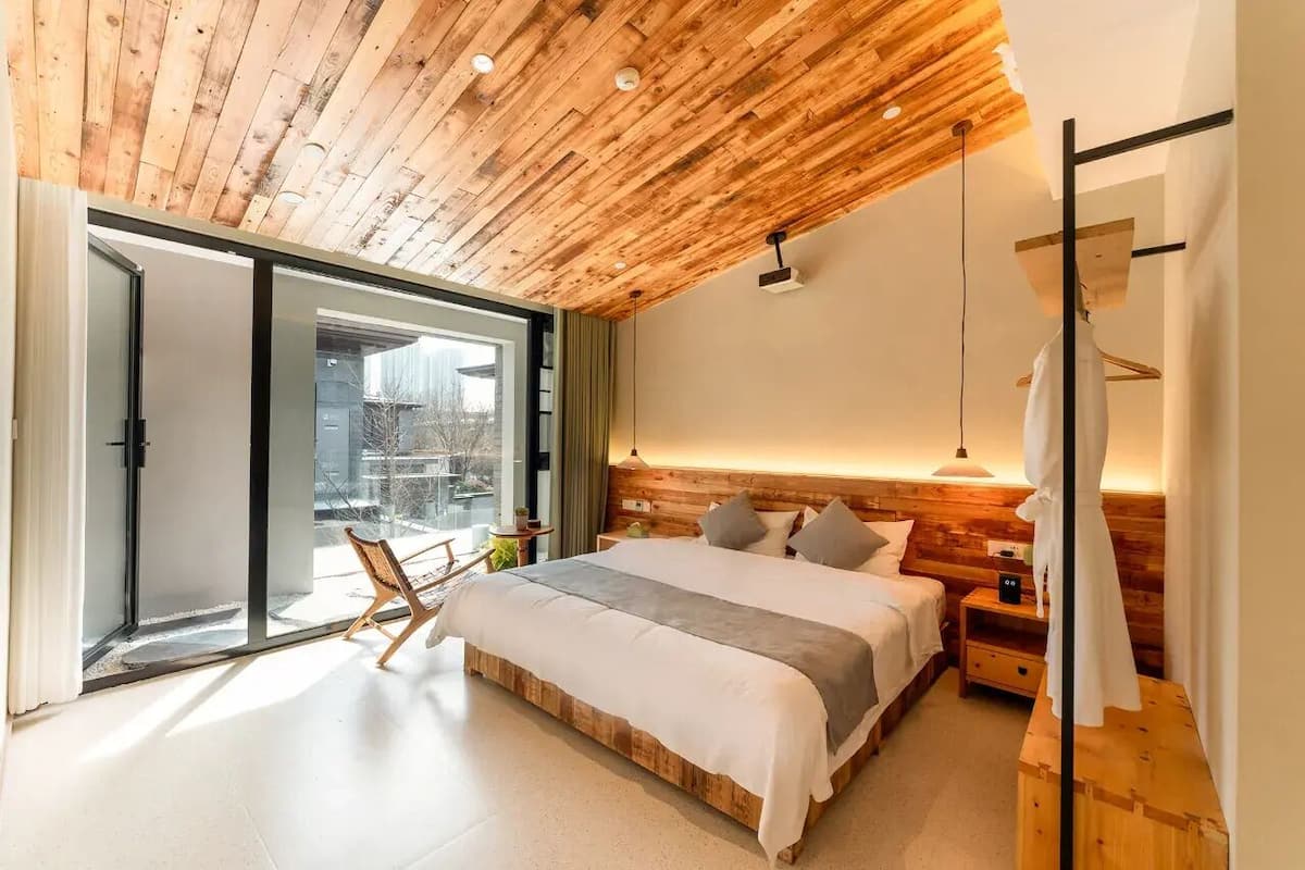 Quarto moderno com teto de madeira, uma cama grande, mesa de cabeceira e vista através de uma porta de vidro deslizante.