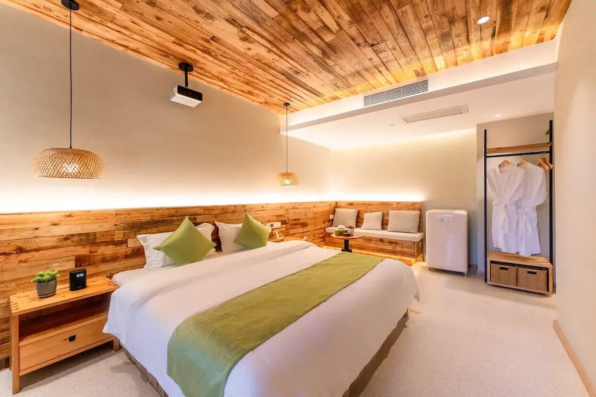 Quarto moderno com cabeceira de madeira, cama com roupa de cama branca, manta verde, mesas de cabeceira com uma planta e um telefone, luminária pendente, pequena mesa com uma cesta de frutas, guarda-roupa branco e roupão branco pendurado na porta.