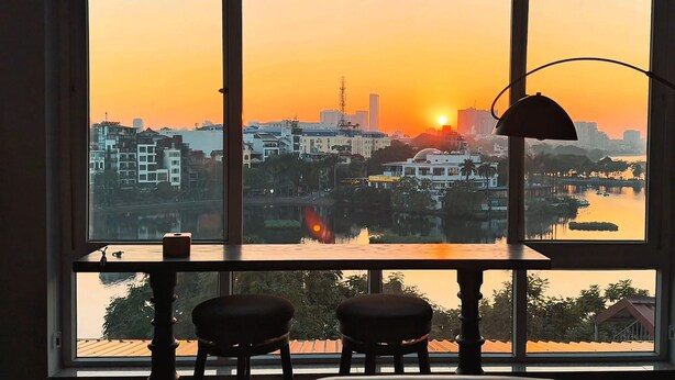A room with a large window overlooking a cityscape at sunset, featuring a table with two chairs and a lamp. - Home. Homestay (Hanoi)