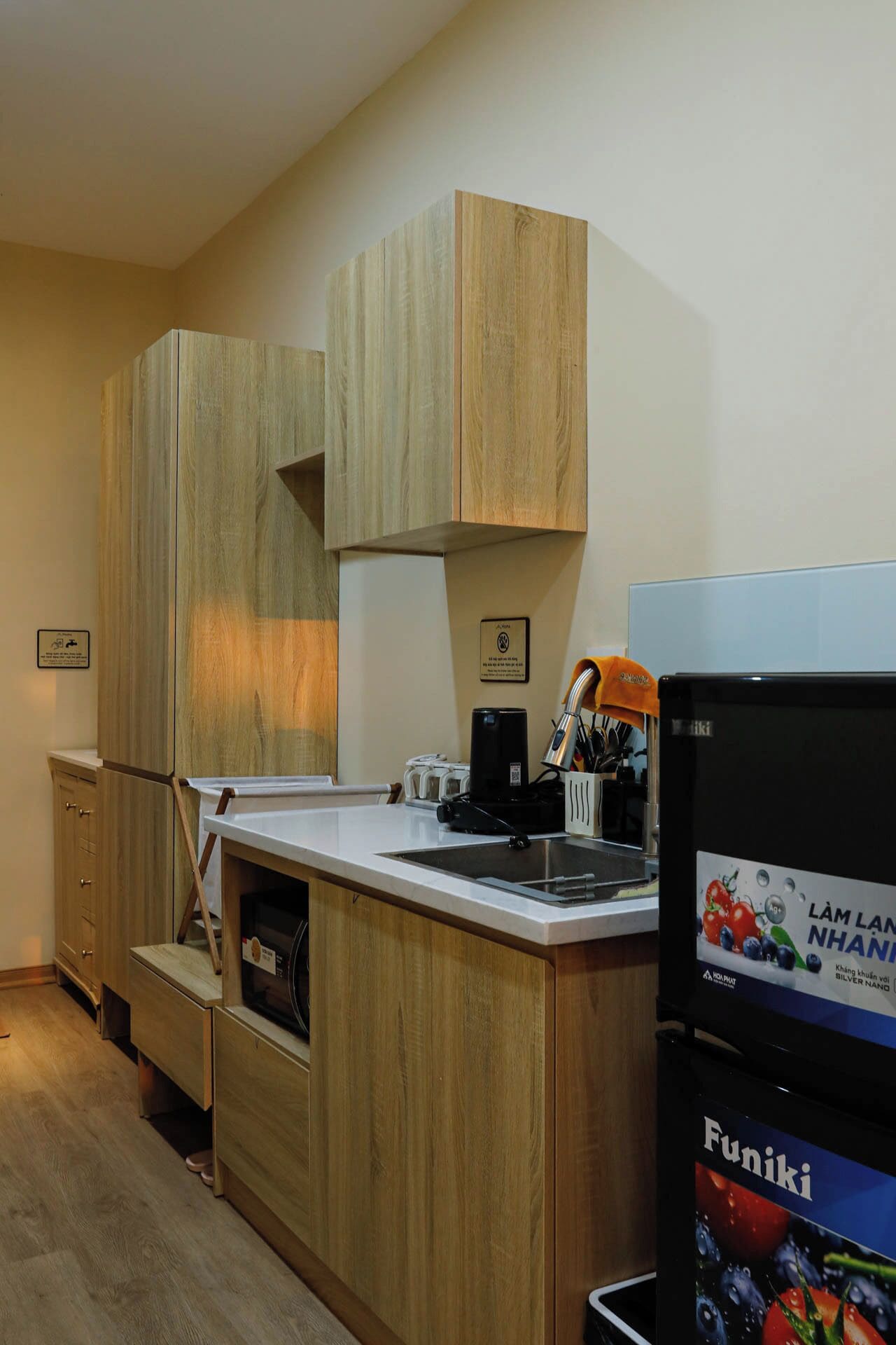 A compact kitchen with wooden cabinets, a built-in oven, a coffee maker, and a refrigerator with a fruit display.