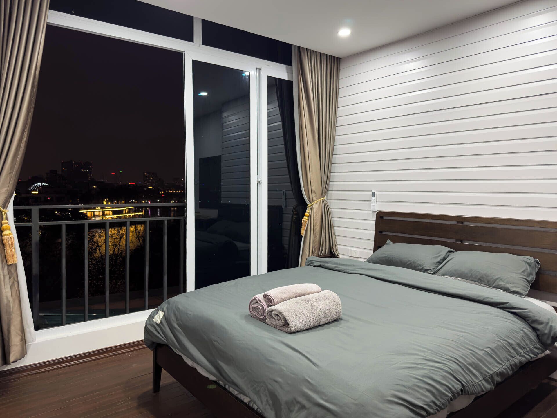Un dormitorio con un ventanal grande, un balcón y vistas nocturnas de la ciudad.