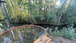 A wooden hot tub with a wooden bench and three glasses, surrounded by a forested area with a river. - Oak Cabin - Hot Tub, BBQ, WiFi & Riverside (Huiscapi)