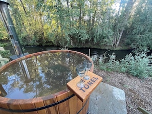 Un bain à remous en bois, avec un banc en bois et trois verres, entouré d’une zone boisée traversée par une rivière.