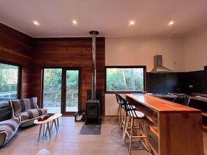 L’intérieur d’un chalet moderne est agrémenté d’un poêle à bois, d’un coin repas avec des tabourets de bar, d’un canapé et de grandes fenêtres offrant une vue sur la forêt.