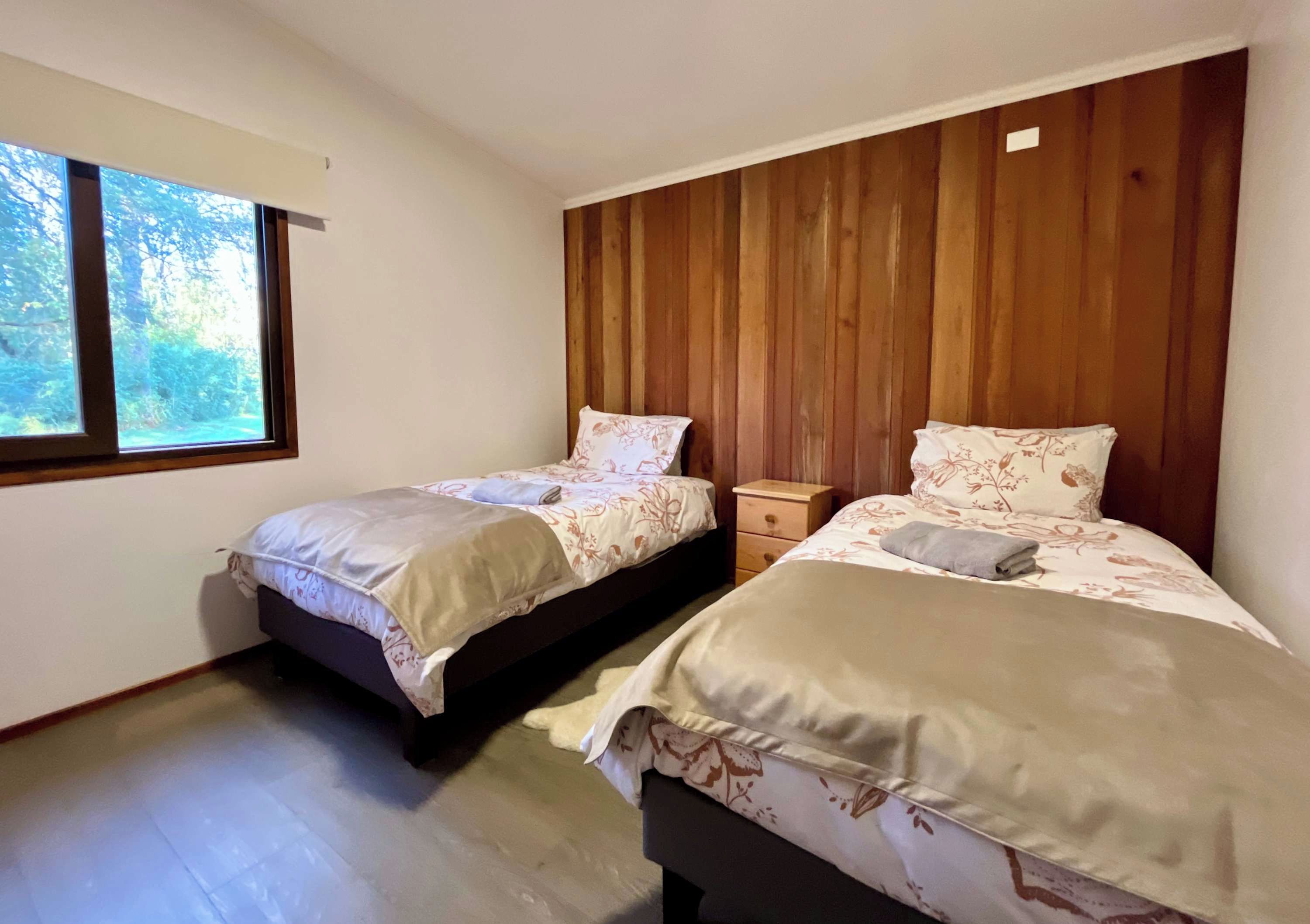 A bedroom with two beds, wooden paneling on the wall, a window, and a nightstand.